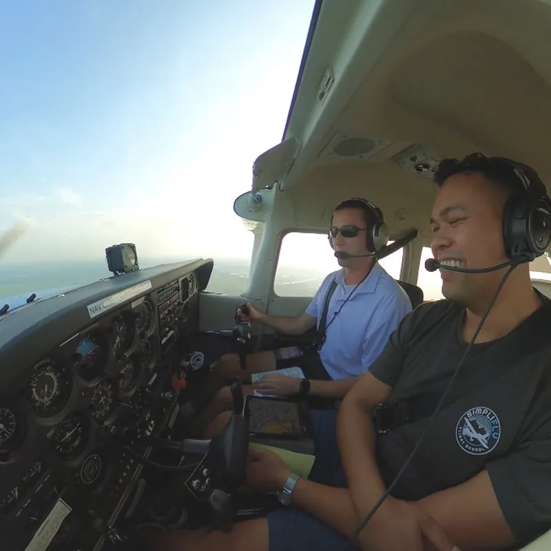 Two pilots in headsets at the controls of a Cessna in cruise, farmland visible through the windshield