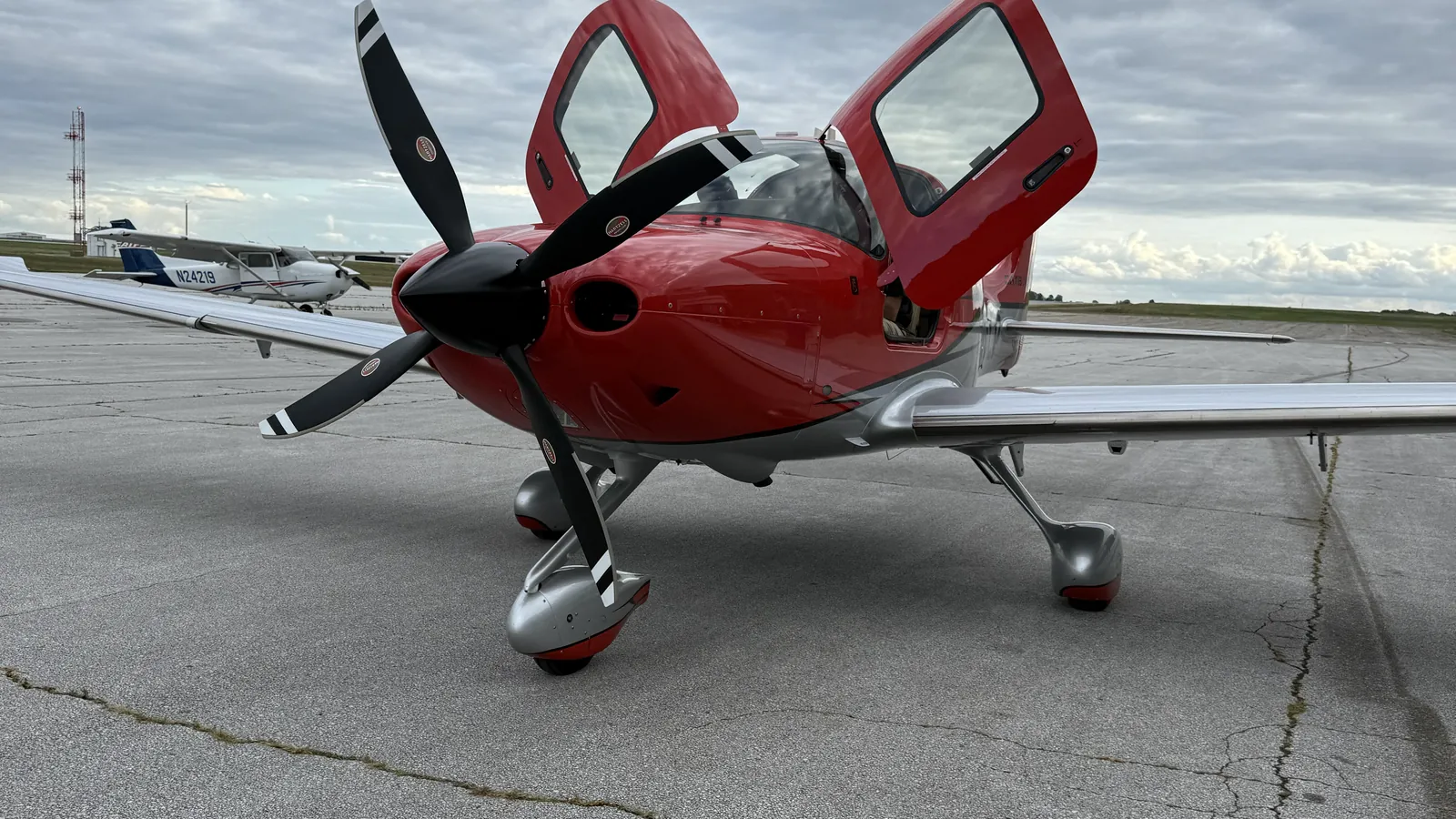 A red Cirrus parked on a concrete ramp with both cabin doors open under a broken cloud layer