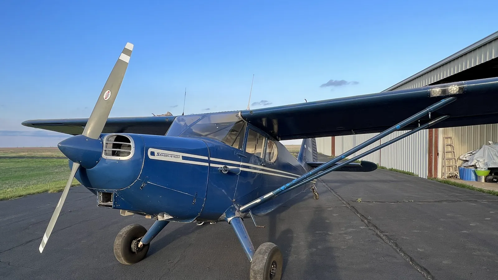 A blue Stinson taildragger parked on a ramp in late afternoon light, hangar and open field behind it