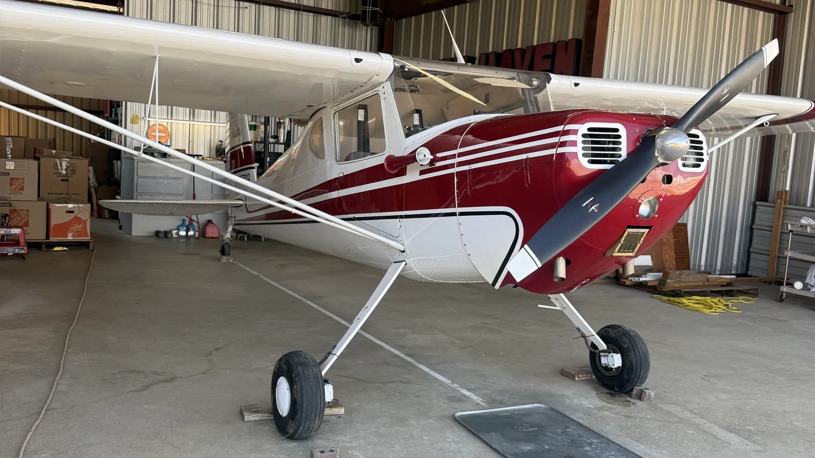 A red and white Cessna taildragger parked inside a metal hangar, chocked on its mains with the tail low and the wing struts in view