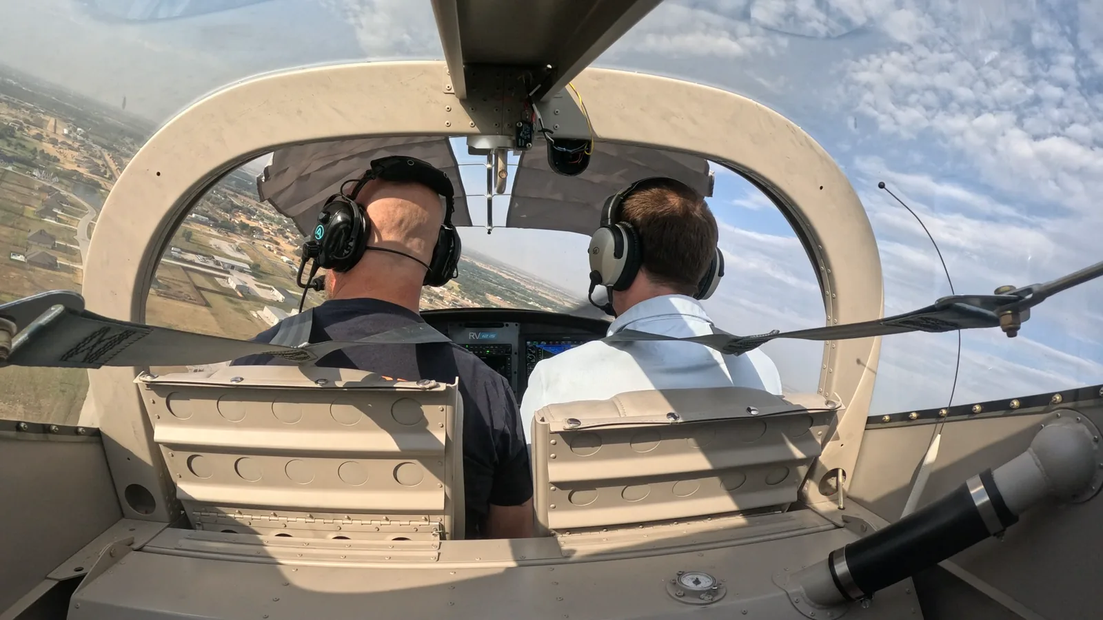 Two pilots side by side in a light airplane cockpit, both in headsets, flying over a city with scattered cloud above